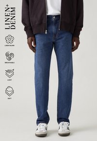 Homme portant un jean en denim mélange lin bleu, une veste noire, une chemise blanche et des baskets blanches sur un fond neutre avec des icônes des qualités du tissu.