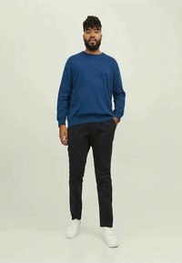 Blauer Pullover mit Rundhalsausschnitt, schwarze Hose und weiße Schuhe. Das Modell steht aufrecht, die Hände in den Taschen, vor neutralem Hintergrund.