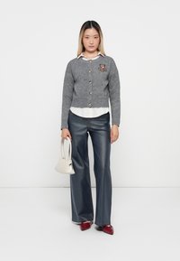 Cardigan grigio lavorato a maglia con bottoni, con ricamo di un orsetto. Abbinato a una camicia bianca con colletto e jeans a gamba larga.