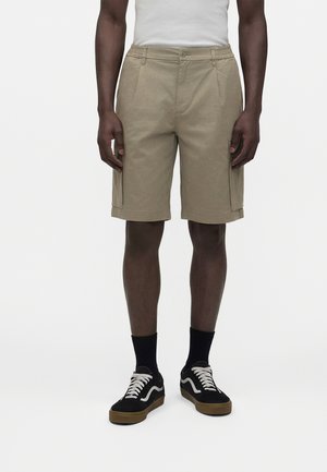 Mann trägt beige knielange Cargo-Shorts, schwarze Socken und schwarze Turnschuhe mit weißen Schnürsenkeln und Sohlen.