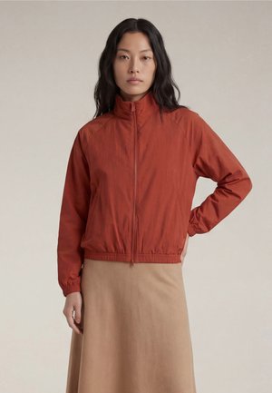 Vrouw met lang donker haar die een roestkleurige jas met rits en een beige rok draagt, staand met één hand op haar heup tegen een effen achtergrond.