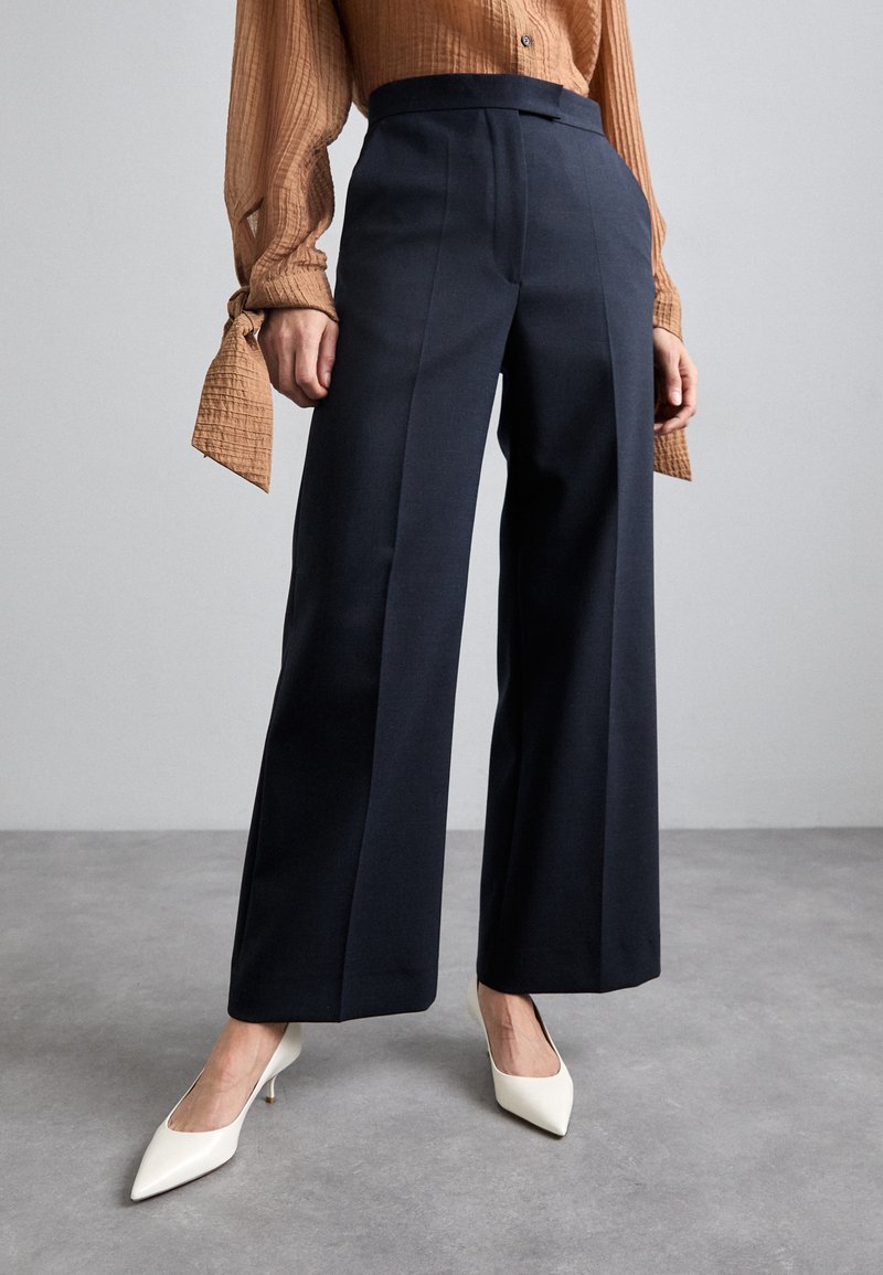 Mørkeblå brede bukser med foranlæg, lavet af glat stof. Matchet med hvide spidse hæle og en brun struktureret skjorte.