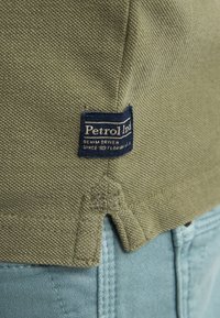 Tissu tissé vert olive avec une étiquette de marque. Présente des détails de couture et une fente à l'ourlet. Assorti avec un denim bleu clair.