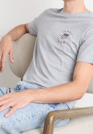 Homme assis sur une chaise beige, portant un t-shirt gris clair GANT 1949 et un jean bleu clair, le bras reposant sur l'accoudoir en bois de la chaise.
