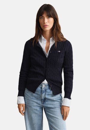 Kvinde iført marineblå kabelstrikket cardigan over hvid skjorte og lyseblå jeans, stående mod en ensfarvet hvid baggrund.