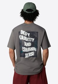 Šedé bavlněné tričko s grafikou na zadní straně, která obsahuje tučný bílý text "VYHÝBEJTE SE GRAVITACI A ZDRAVÉMU ROZUMU" a dekorativní akcenty.
