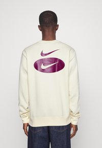 Krémová mikina s velkými fialovými logy Nike na zádech, žebrovanými manžetami a spodním lemem. Měkká tkanina s uvolněným střihem.