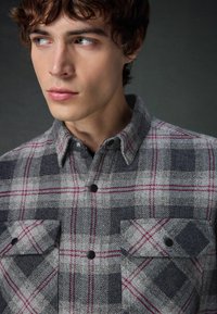 Cămașă flanelată în carouri gri și bleumarin cu accente roșii, design cu nasturi și două buzunare frontale. Material texturat și guler ascuțit.
