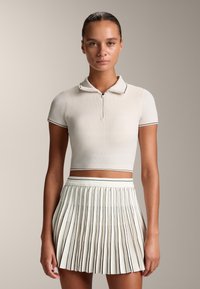 Ljust beige kortärmad polo med dragkedja, kombinerad med en vit plisserad kjol med tunna svarta ränder vid fållen.