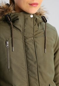 Zelená zateplená bunda s kapucí lemovanou umělou kožešinou, se šňůrkami a stříbrným zipem. Hladký povrch s prošívaným vzorem.