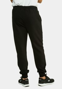 Schwarze Jogginghose mit elastischem Bund und Bündchen an den Knöcheln, aus weichem Stoff. Kombiniert mit schwarzen Sportschuhen mit weißen Sohlen.
