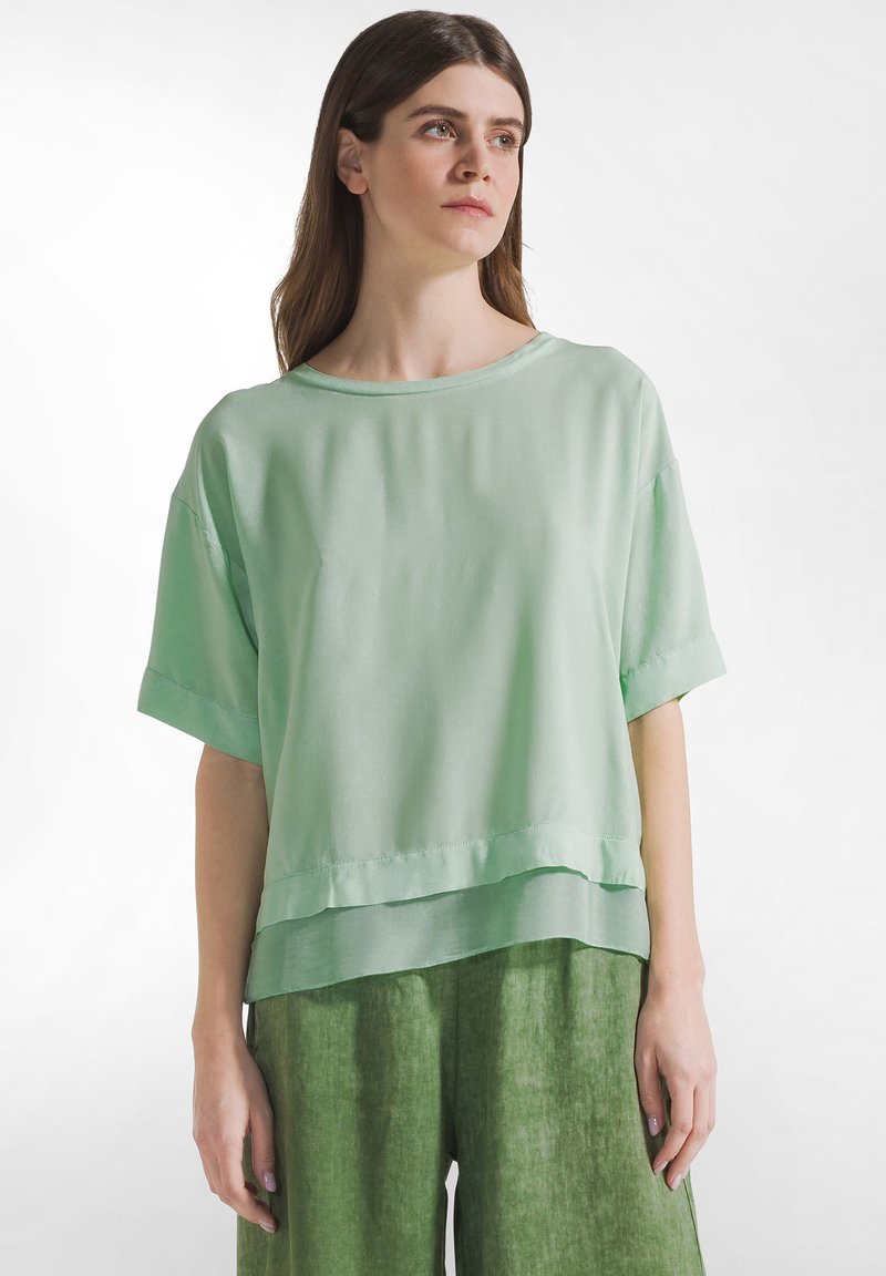 Kvinde iført en løs, lysegrøn top med korte ærmer og mørkere grønne bukser, stående mod en ensfarvet hvid baggrund, kiggende en smule mod højre.