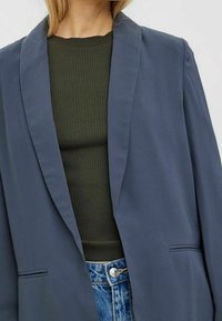 Marinblå blazer med sjalkrage, tillverkad av slät tyg. Kombinerad med en ribbad mörkgrön topp och ljusblå denimjeans.