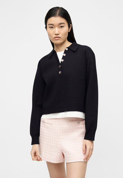 Γυναίκα που φοράει μαύρο μακρυμάνικο πουκάμισο πόλο με κουμπιά πάνω από λευκό τοπ, συνδυασμένο με ροζ ανοιχτόχρωμο σορτς με υφή και διακοσμητικές τσέπες.