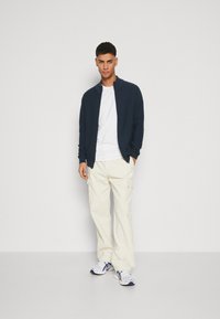 Sudadera navy con cremallera y textura acanalada, camiseta blanca por debajo, pantalones cargo color crema y zapatillas blancas con detalles azules.