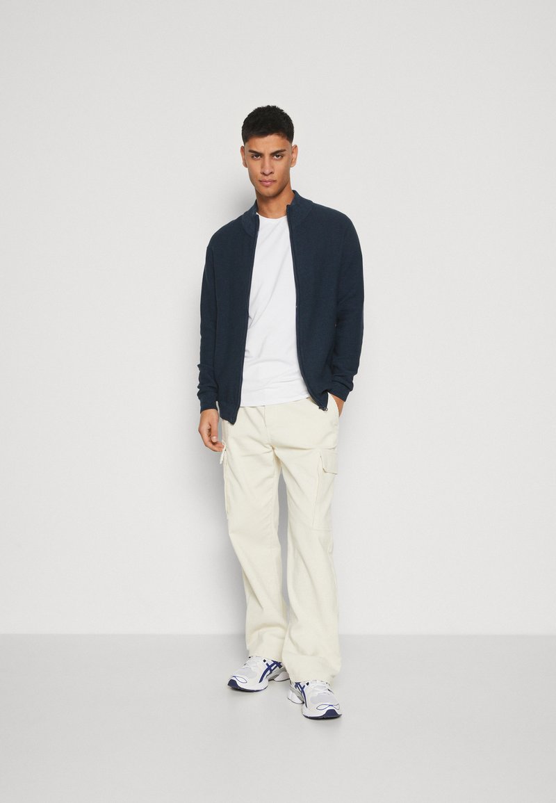 Granatowy sweter na zamek błyskawiczny o prążkowanej fakturze, biała koszulka pod spodem, kremowe spodnie cargo oraz białe sneakersy z niebieskimi akcentami.