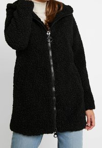 Giacca in pile nera con superficie texturizzata e soffice, con zip frontale e cappuccio. Taglio a metà coscia, presenta maniche lunghe e una vestibilità rilassata.