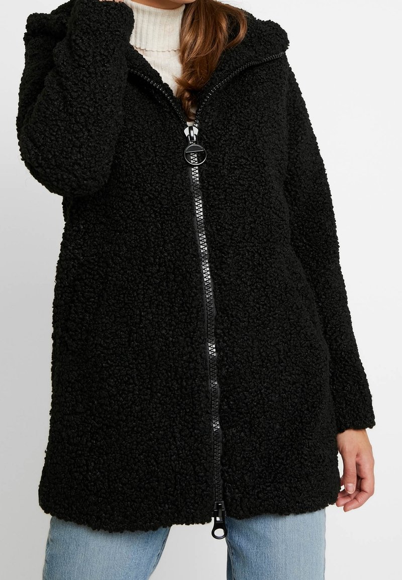 Giacca in pile nera con superficie texturizzata e soffice, con zip frontale e cappuccio. Taglio a metà coscia, presenta maniche lunghe e una vestibilità rilassata.