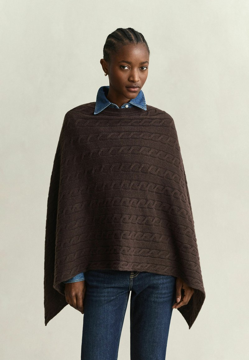 Femme aux cheveux tressés portant un poncho marron texturé par-dessus une chemise en denim et un jean bleu, debout devant un fond clair uni.