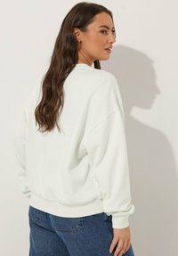 Sudadera blanca de gran tamaño con costuras de hombro caídas y puños acanalados. Combinada con jeans de mezclilla de cintura alta. Textura de algodón suave.