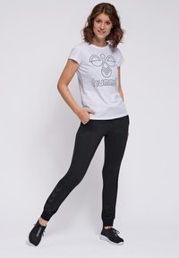 Weißes T-Shirt mit einem schwarzen geometrischen Logo und Text, kombiniert mit schwarzen engen Hosen, die an der Seite ein Zickzackmuster aufweisen. Schwarze Sportschuhe.