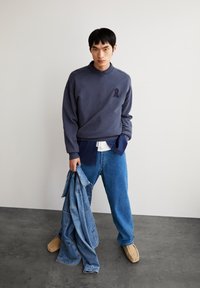 Sweat-shirt bleu marine avec un petit emblème sombre, superposé sur une chemise bleu marine, associé à un jean bleu clair et des mocassins beiges, tenant une veste en denim.
