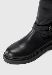 Stivale alla caviglia in pelle nera con punta tondeggiante, dettagli cuciti e fascia laterale decorativa con fibbia in metallo su uno sfondo bianco.