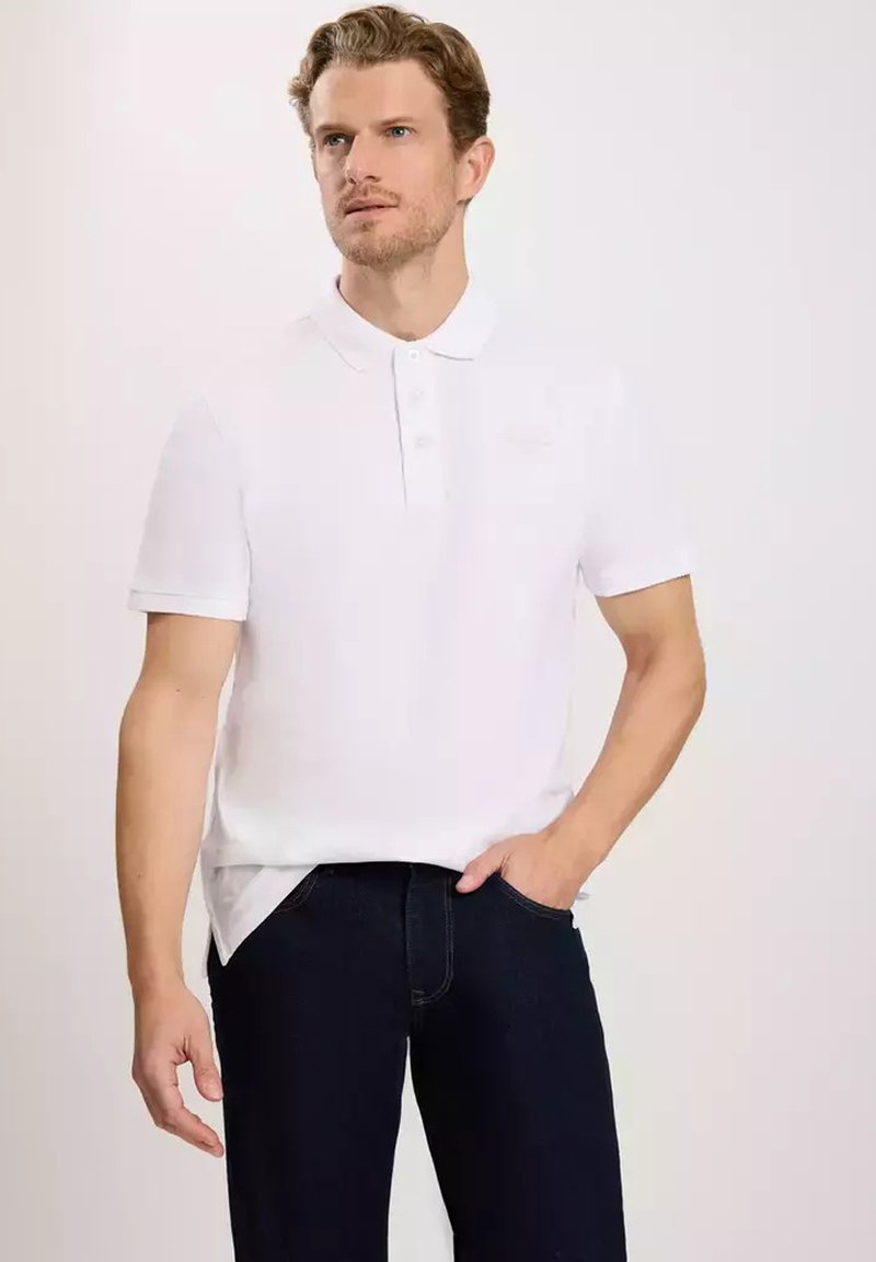 Biała koszulka polo z klasycznym kołnierzykiem i trzyguzikowym zapięciem. Gładka bawełniana tkanina, krótkie rękawy, w zestawie z ciemnymi dżinsami.