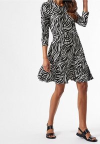 Vestido de rayas de cebra en blanco y negro, con mangas de tres cuartos, cintura entallada, falda acampanada y dobladillo con volantes. Lleva sandalias negras.