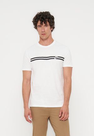 Mand med krøllet mørkt hår iført en hvid t-shirt med korte ærmer med sorte striber og beige bukser, stående mod en ensfarvet hvid baggrund.