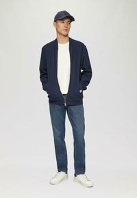 Navy zip-up jacket over een wit shirt, gecombineerd met blauwe jeans en witte sneakers. De jas heeft een kraagloos ontwerp.