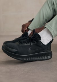 Sorte sportsko med mesh-overdel, tekstureret sål og tonet Nike-logo. Snørebåndene er sorte, og der er kontrastdetalje i hvid ved sokken.