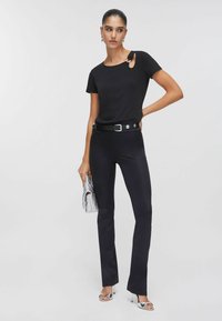 Schwarzes kurzärmeliges Oberteil mit seitlichem Ausschnitt, kombiniert mit eng anliegenden schwarzen Hosen, einem schwarzen Gürtel mit silbernen Details und silbernen Sandalen mit hohen Absätzen.