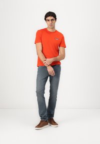 Oranžinis trumparankovis marškinėlis su logotipu kairėje krūtinės pusėje, derinamas su siaurėjantys mėlyni džinsais ir rudi sportbačiai. Paprasta fono.