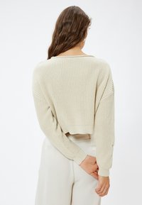 Beige gebreide trui met een cropped ontwerp en losse mouwen, voorzien van een textuurpatroon. De achterkant heeft een brede halslijn en een ribgebreide zoom.