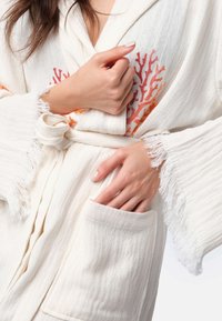 Biela róba vyrobená z textúrovanej tkaniny, s rukávmi s strapcami a koralovým vzorom v oranžovej a červenej farbe. Obsahuje viazaný opasok a bočnú kapsu.