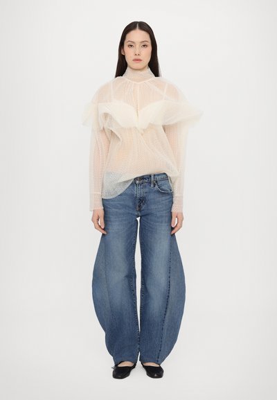 Διαφανής λευκή μπλούζα με διχτυωτό σχέδιο και βολάν, συνδυασμένη με φαρδιά μπλε τζιν. Το σύνολο έχει χαλαρή εφαρμογή και μοντέρνο σχέδιο.