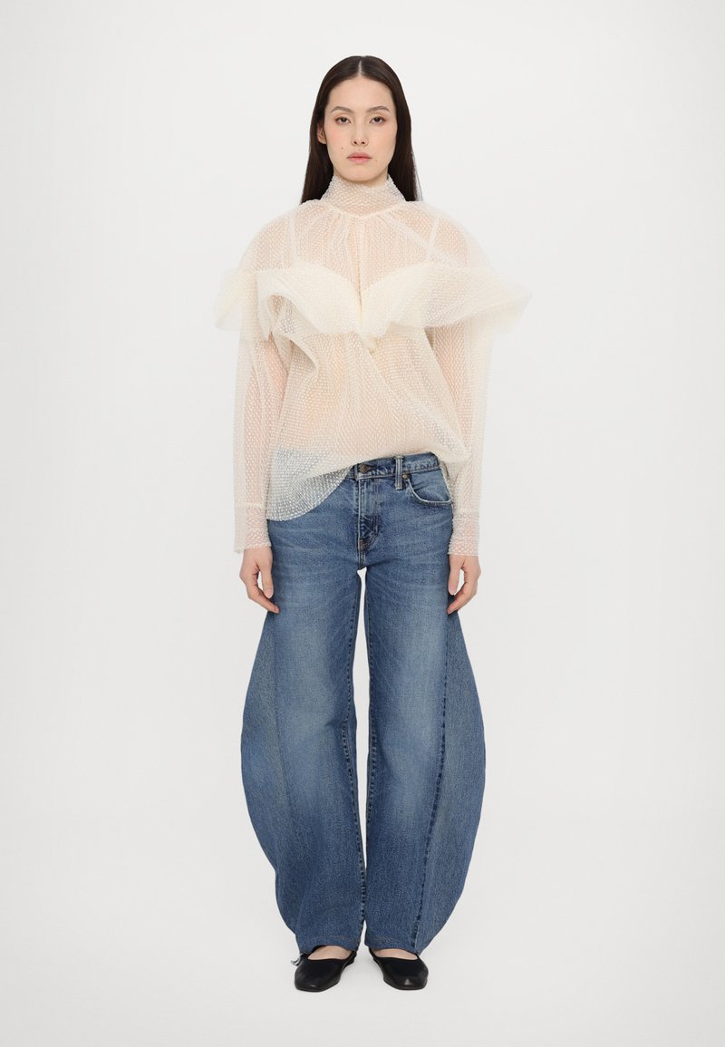 Gennemsigtig hvid mesh-top med flæsedetaljer, parret med brede ben blå jeans. Outfittet har en afslappet pasform og et moderne design.