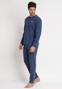 Maglietta a maniche lunghe blu navy con piping bianco, abbinata a pantaloni da pigiama a quadri blu. Materiale misto cotone, vestibilità comoda e branding minimale.