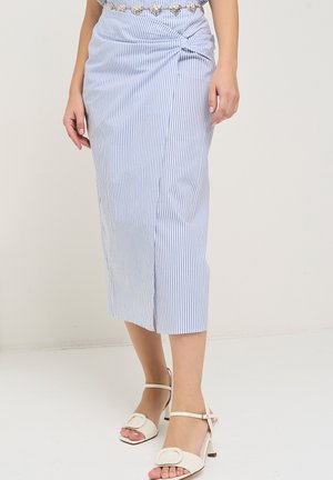 Vrouw draagt een blauw-wit gestreepte midirok met een geplooide knoop aan de zijkant en witte open teen sandalen met hak en enkellijnen.