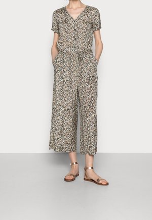 Femme portant une combinaison short manches courtes à motif floral avec un pantalon large raccourci et des sandales plates métalliques à bride cheville, mains dans les poches.