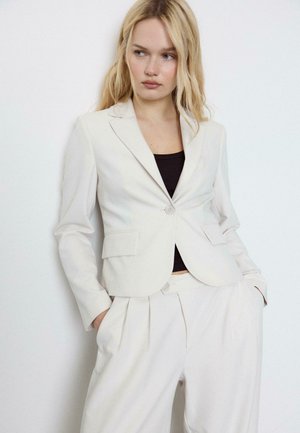 Blonde Frau, die einen weißen Blazer und passende hoch taillierte Hosen trägt, mit Händen in den Taschen, seitlich blickend vor weißem Hintergrund.