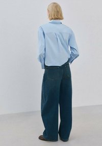 Cămașă albastru deschis cu nasturi, cu mâneci lungi, asortată cu blugi evazați din denim închis. Are pliuri la spate și două buzunare în partea din spate.