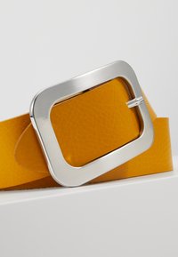 Žltý kožený opasok so štvorcovou striebornou prackou. Koža má textúrovaný vzor a pracka má elegantný, moderný dizajn.