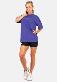 Paarse oversized t-shirt met korte mouwen en geborduurd logo, gecombineerd met zwarte sportshorts en witte sneakers.