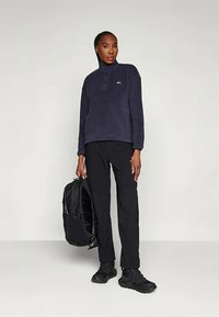 Person wearing navy pullover, black pants, and black shoes, holding a black backpack against a plain light background.