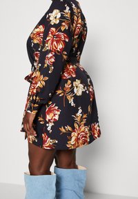 Květinové šaty s tmavě námořnickou tkaninou a červenými a žlutými růžemi, s úzkým pasem, dlouhými rukávy a délkou do kolen. Spárované se světle modrými botami.