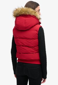 Rote Steppweste mit einem fellgefütterten Kapuze, die über gesteppten Bereichen verfügt. Die Ärmel sind schwarz mit einem Zopfmuster. Rückansicht, getragen über einem roten Oberteil.