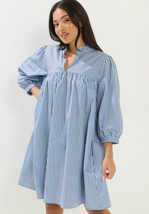 Młoda kobieta z długimi ciemnymi włosami, ubrana w luźną sukienkę w pionowe niebiesko-białe paski z trójkątnymi bufiastymi rękawami do połowy ramienia.