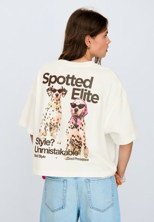 Kobieta ubrana w białą oversize koszulkę z dwoma cętkowanymi psami w okularach przeciwsłonecznych i napisem "Spotted Elite, Style? Unmistakable" na plecach.